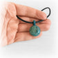 Hand holding a memorial pendant hand crafted from teal and aqua glass on a black leather chord