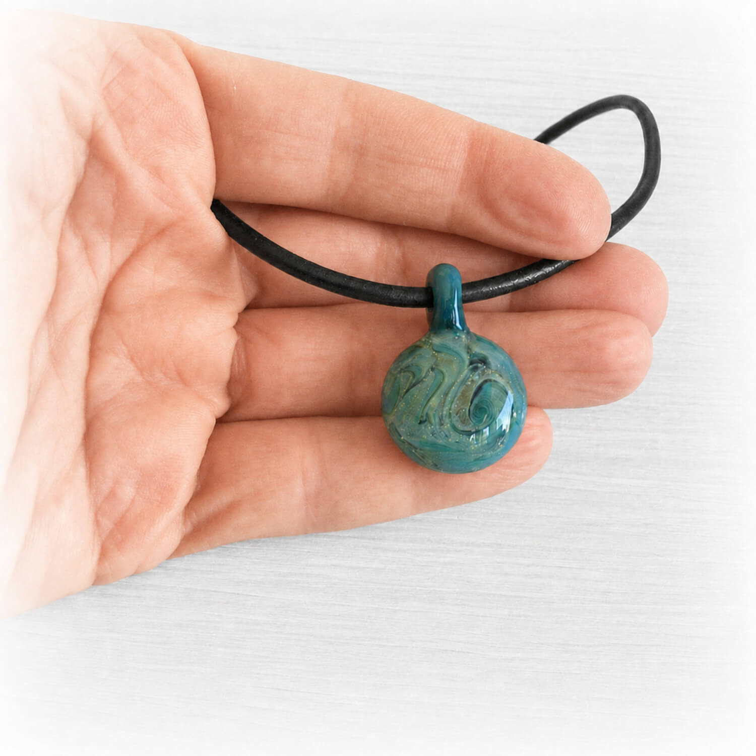 Hand holding a memorial pendant hand crafted from teal and aqua glass on a black leather chord