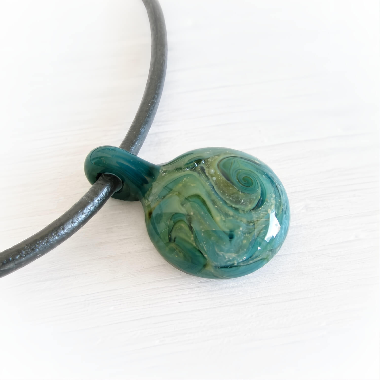 Hand blown glass cremation pendant with swirls of teal, aqua and cremation ashes on a black leather chord