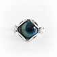A silver ring featuring a unique hexagonal setting with a swirling blue, green, and black cremation glass center, photographed on a light, textured white background.