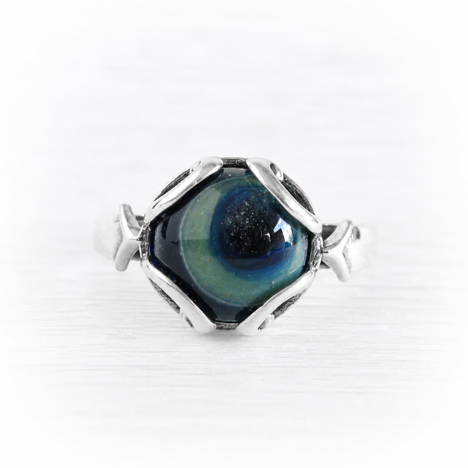 A silver ring featuring a unique hexagonal setting with a swirling blue, green, and black cremation glass center, photographed on a light, textured white background.