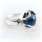 A sterling silver ring featuring a round, polished blue glass cremation cabochon with swirling patterns, set in a decorative silver bezel on a white textured background.