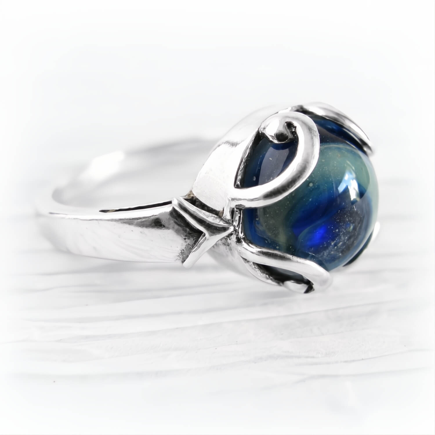 A sterling silver ring featuring a round, polished blue glass cremation cabochon with swirling patterns, set in a decorative silver bezel on a white textured background.