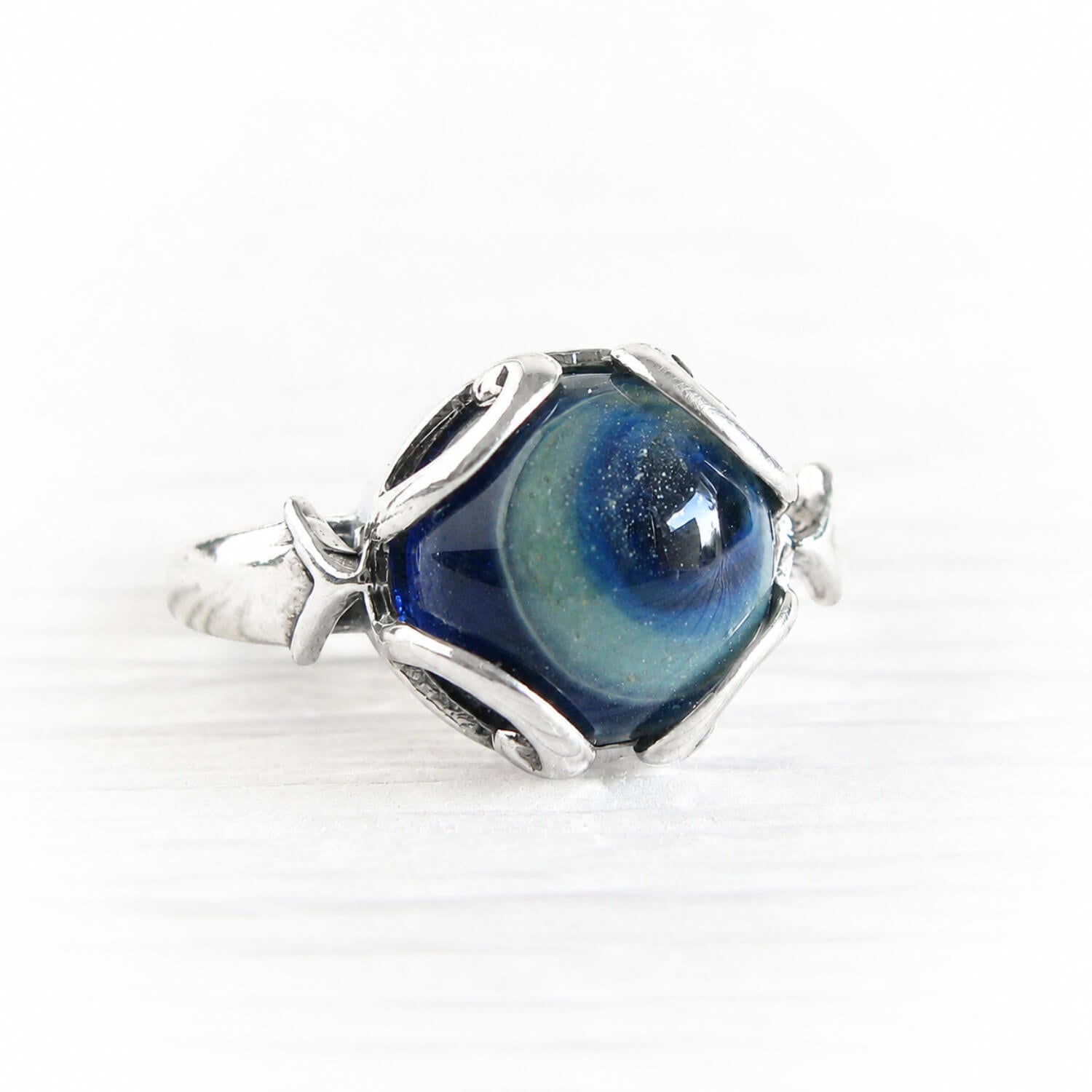 A silver ring featuring a hexagonal setting and cremation glass cabochon with a swirling blue and green marbled pattern, set against a clean white background.