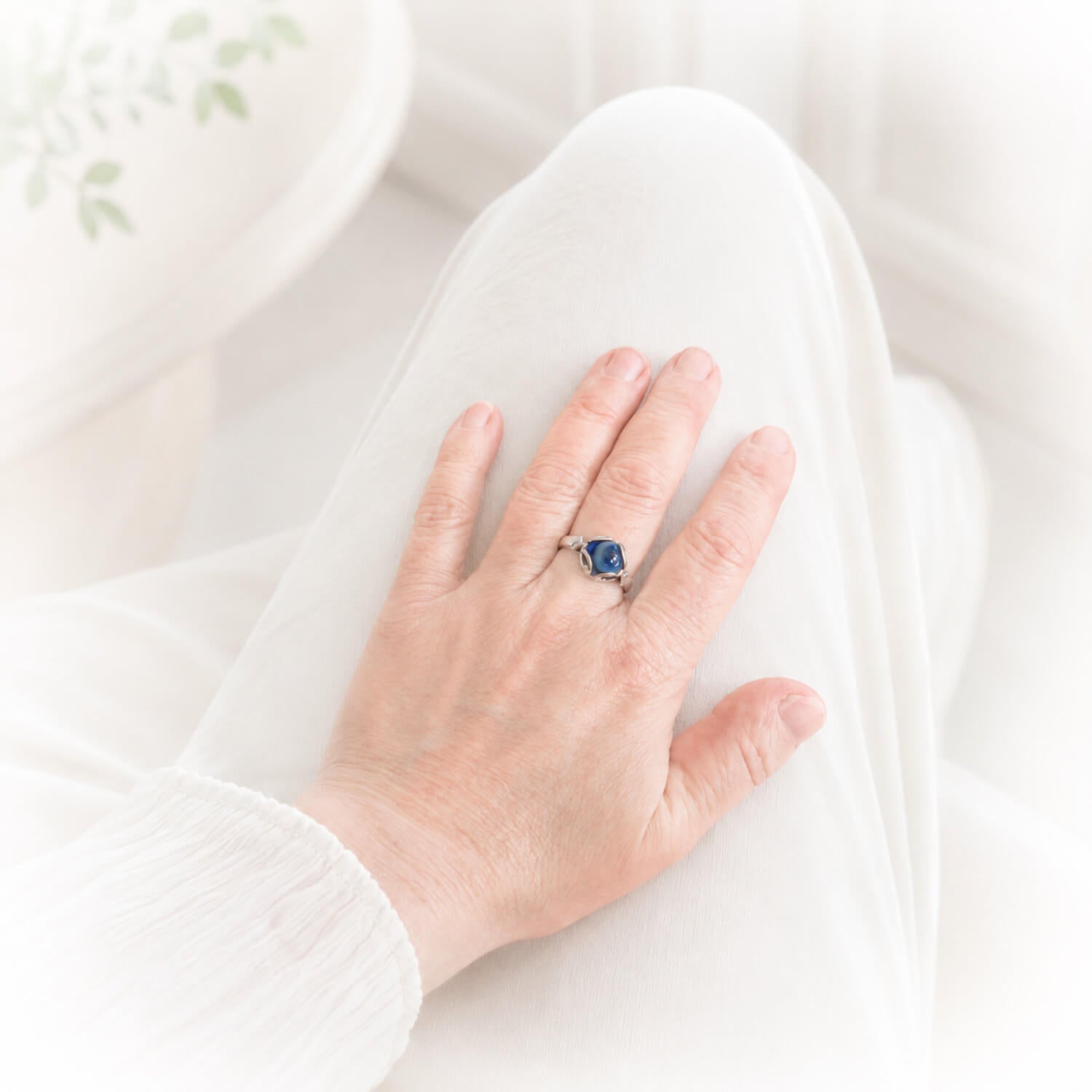 A silver ring with a blue cremation glass cabochon worn on a person's ring finger, shown resting on their lap against a soft white background.
