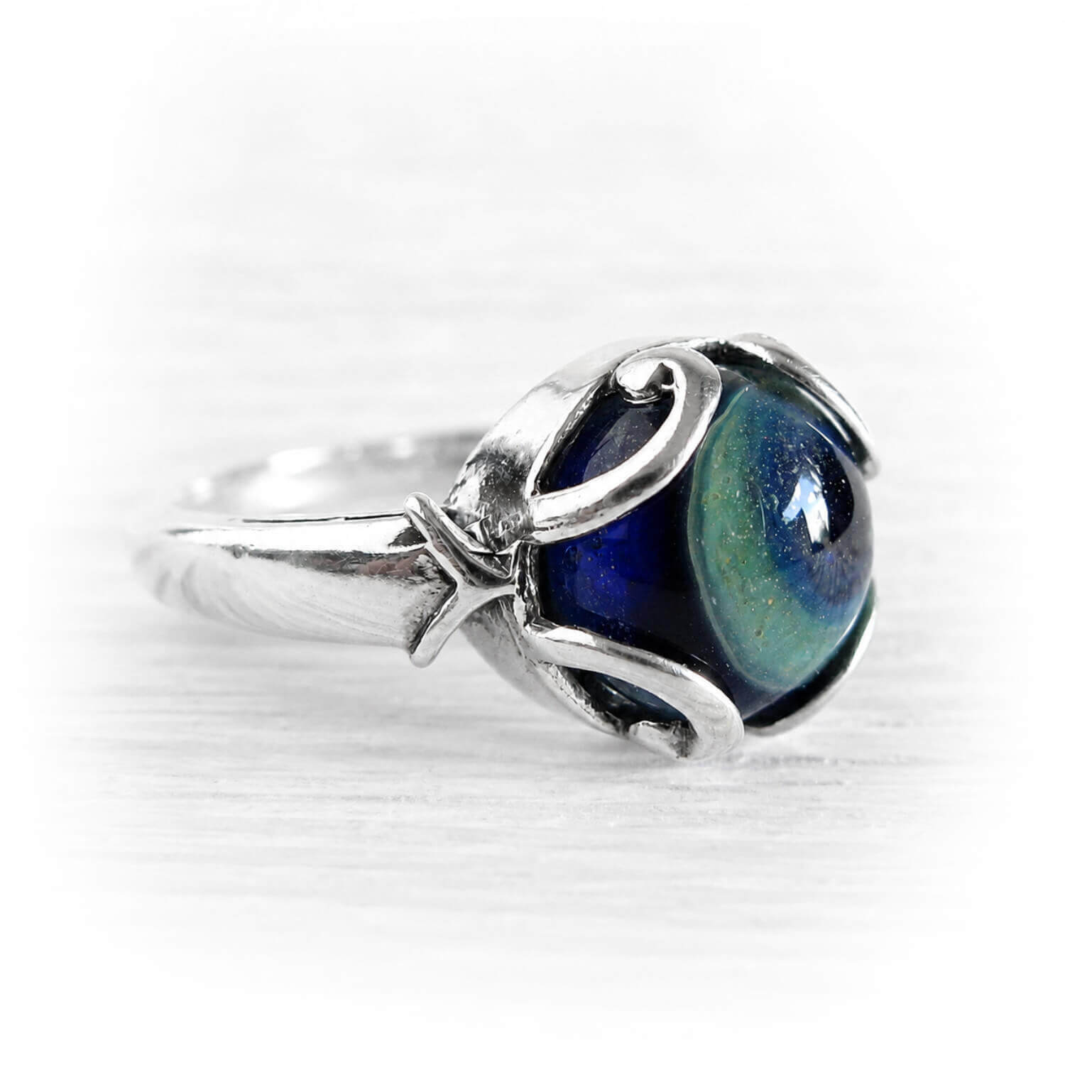 A sterling silver ring featuring a round cremation glass cabochon with a deep blue and green iridescent pattern, set in a decorative swirl design on a white background.