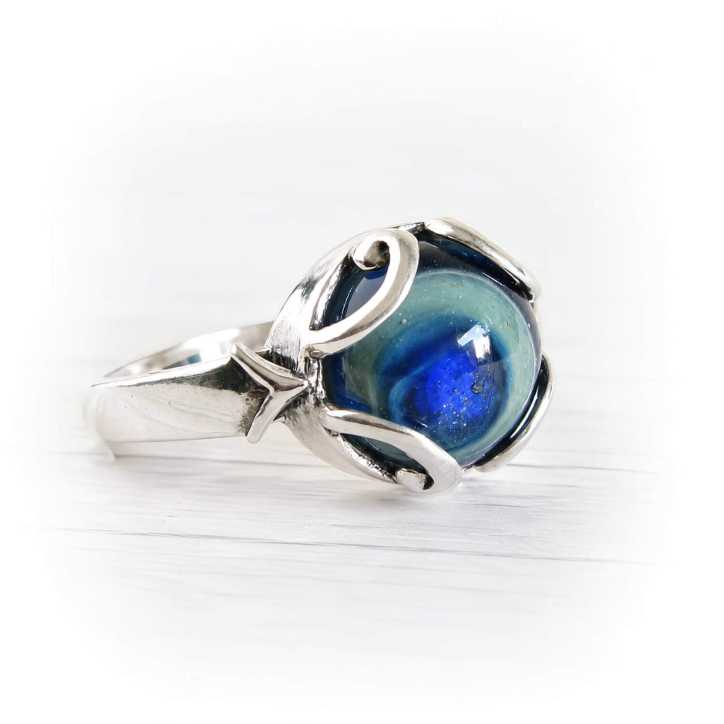 A silver ring featuring a round, marbled blue and green cremation glass cabochon, set in a polished sterling silver band with organic, flowing curves, photographed on a white wooden surface.