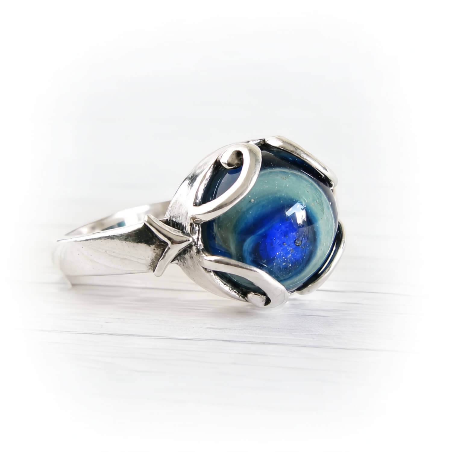 A silver ring featuring a round, marbled blue and green cremation glass cabochon, set in a polished sterling silver band with organic, flowing curves, photographed on a white wooden surface.