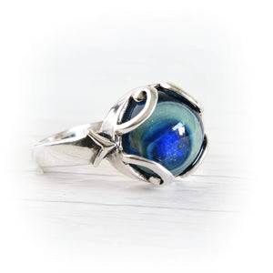 A silver ring featuring a round, marbled blue and green cremation glass cabochon, set in a polished sterling silver band with organic, flowing curves, photographed on a white wooden surface.