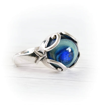 A silver ring featuring a round, marbled blue and green cremation glass cabochon, set in a polished sterling silver band with organic, flowing curves, photographed on a white wooden surface.