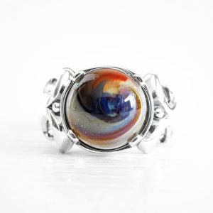 A silver ring featuring a round cabochon cremation glass stone with a swirling marbled pattern of blue, orange, brown, and cream, set against a clean white background.