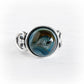 A silver ring featuring a round cabochon cremation glass stone with a swirling blue, brown, and black cosmic or galaxy-like pattern, set against a clean white background.