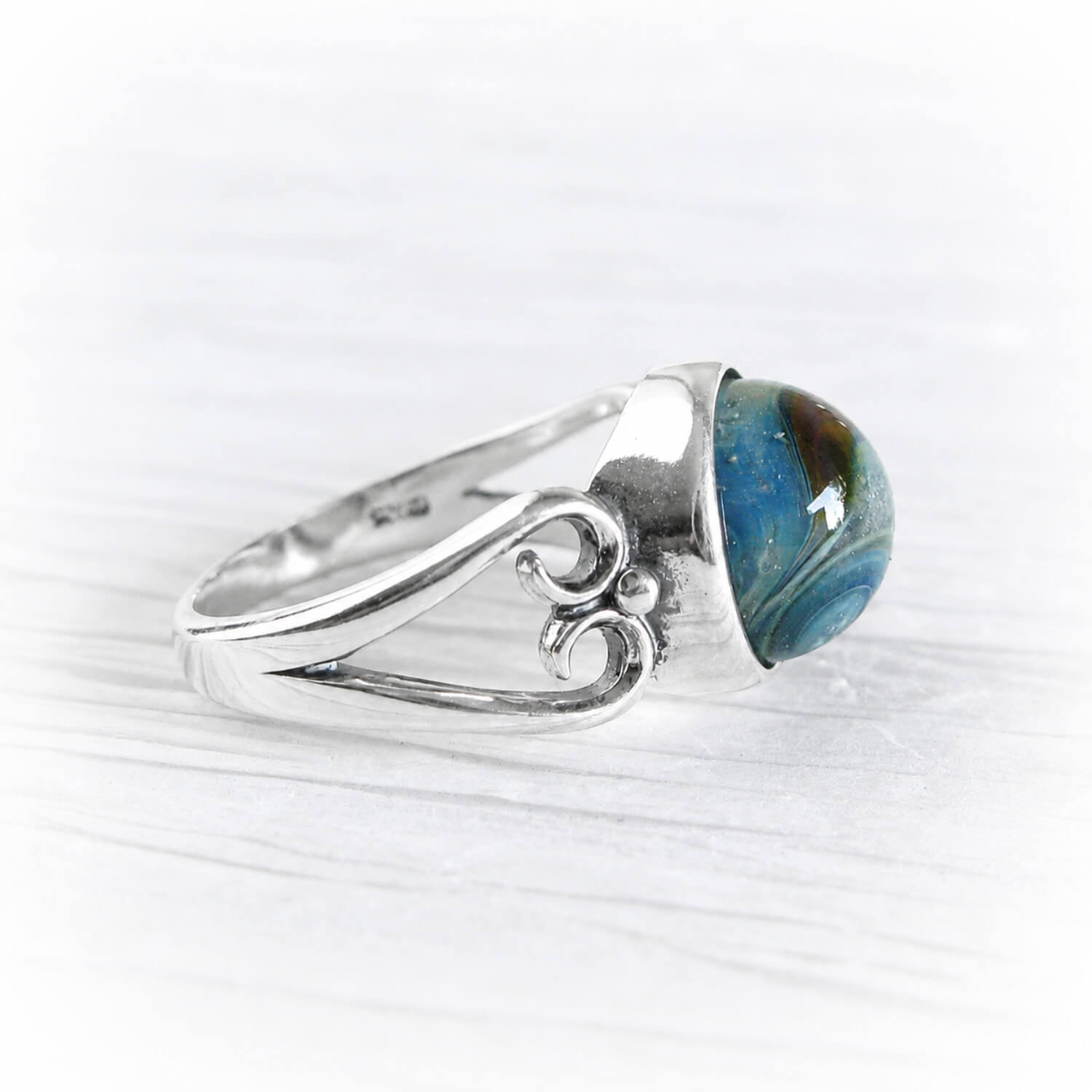 A sterling silver ring featuring a round cremation  glass cabochon gemstone with a marbled blue, green, and brown pattern, displayed on a light gray wood-textured background.