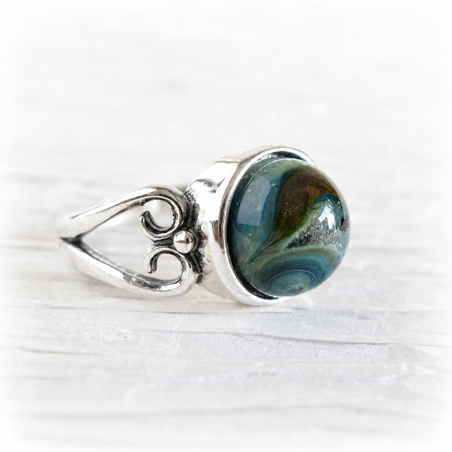 A silver ring featuring a round cremation glass cabochon gemstone with a marbled pattern of teal, blue, and brown, set on a light, textured white background.
