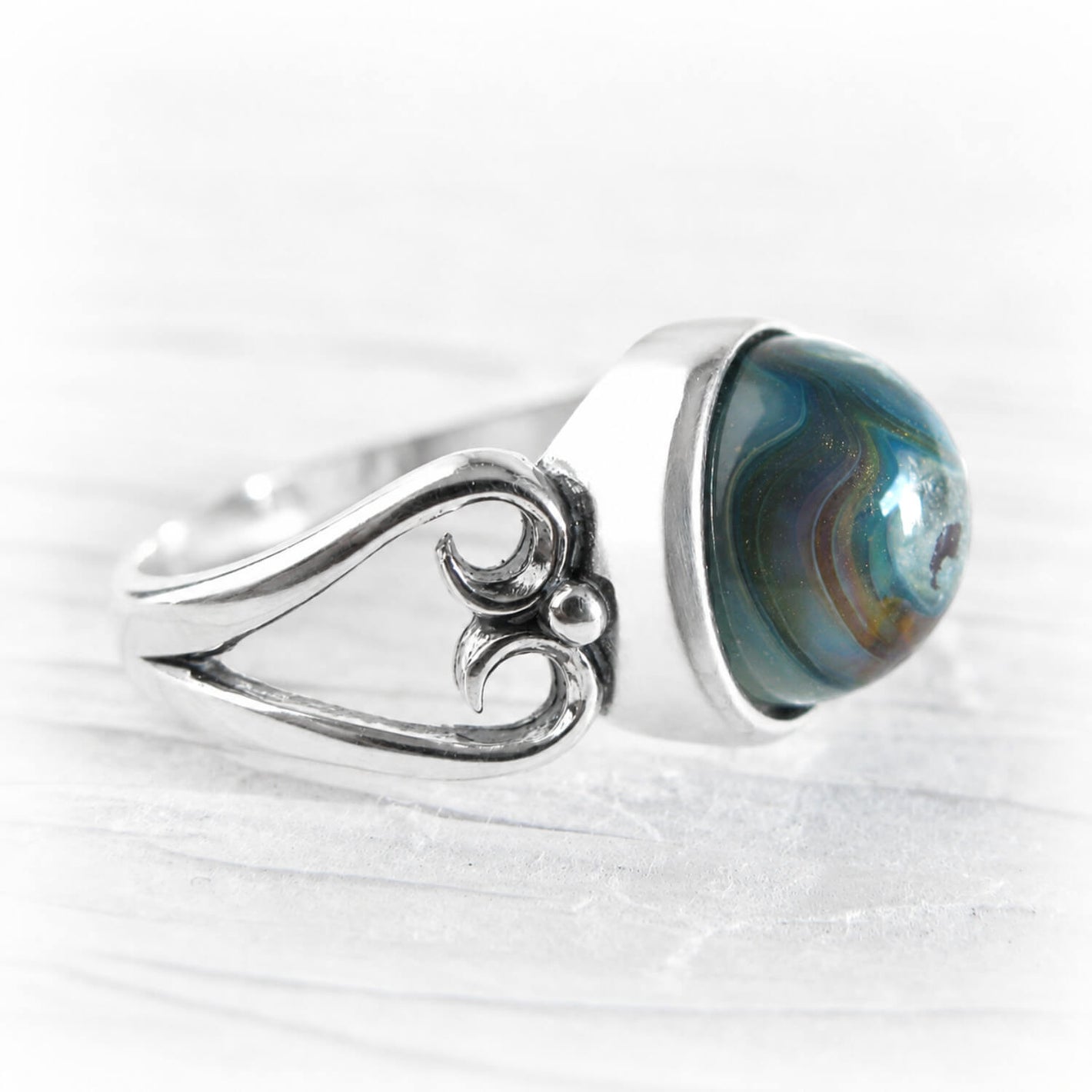 A silver ring featuring a large, round cremation glass cabochon gemstone with swirling blue, green, and brown agate-like patterns, set on a textured white background.
