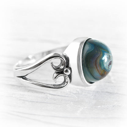 A silver ring featuring a large, round cremation glass cabochon gemstone with swirling blue, green, and brown agate-like patterns, set on a textured white background.
