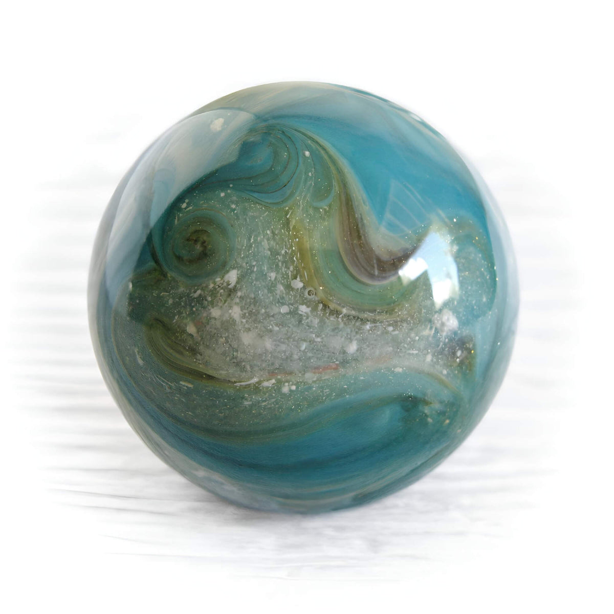A round glass cremation ash marble with a marbled swirl pattern in shades of teal blue, olive green, and white, resting on a white surface.