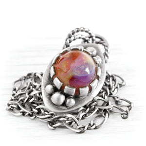 A silver-toned memorial pendant necklace featuring a textured, organic-shaped glass cremation cabochon stone with swirling orange, red, and purple colors, resting on a white background.