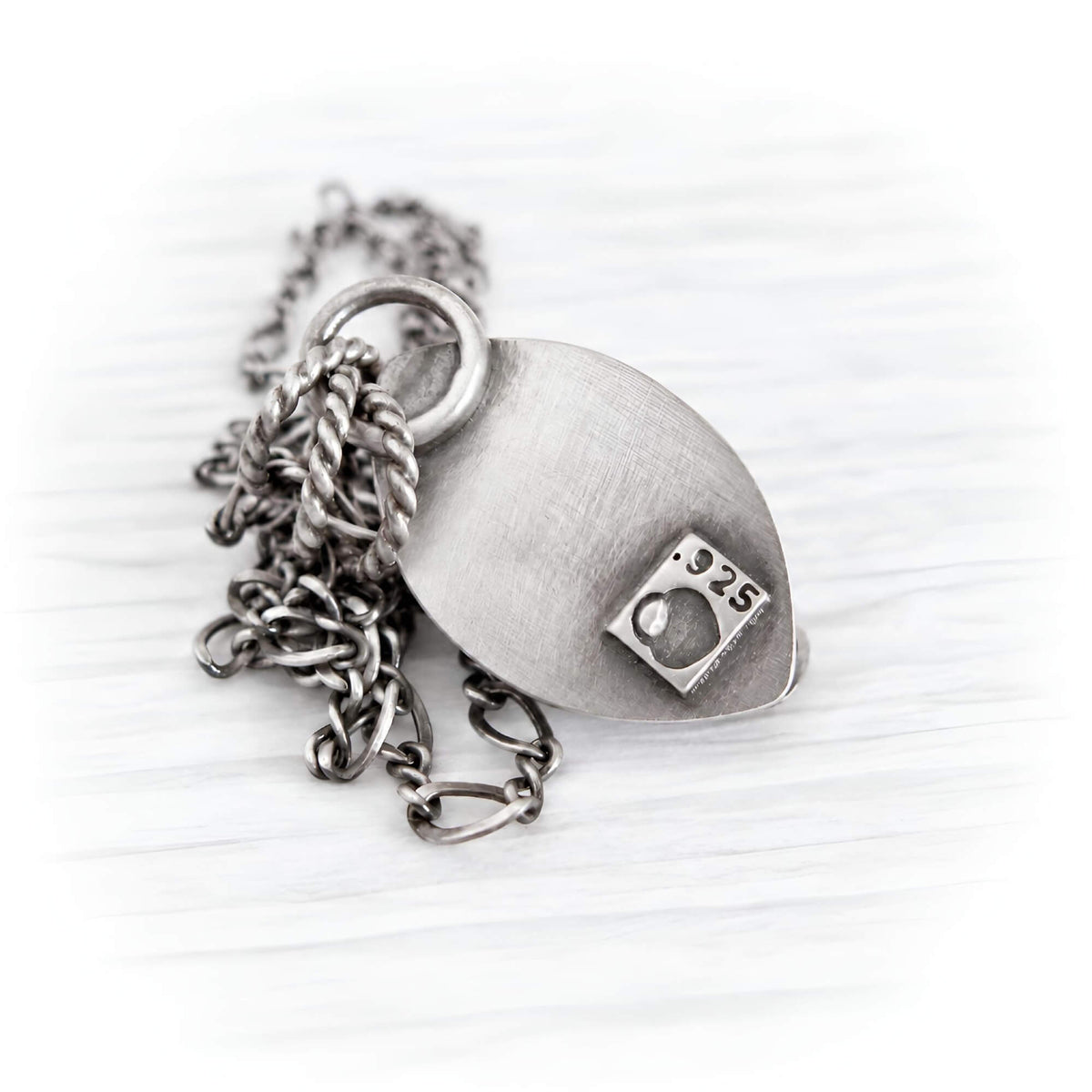 A silver-toned pendant necklace featuring a brushed metal teardrop-shaped tag with a small square engraving that reads '925', attached to a layered chain with a twisted rope link and a curb link, displayed on a light wood background.
