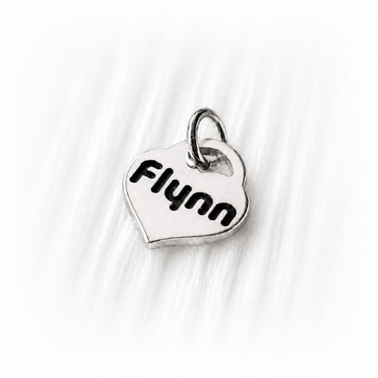 Silver heart-shaped charm with 'Flynn' engraved on a white fabric background
